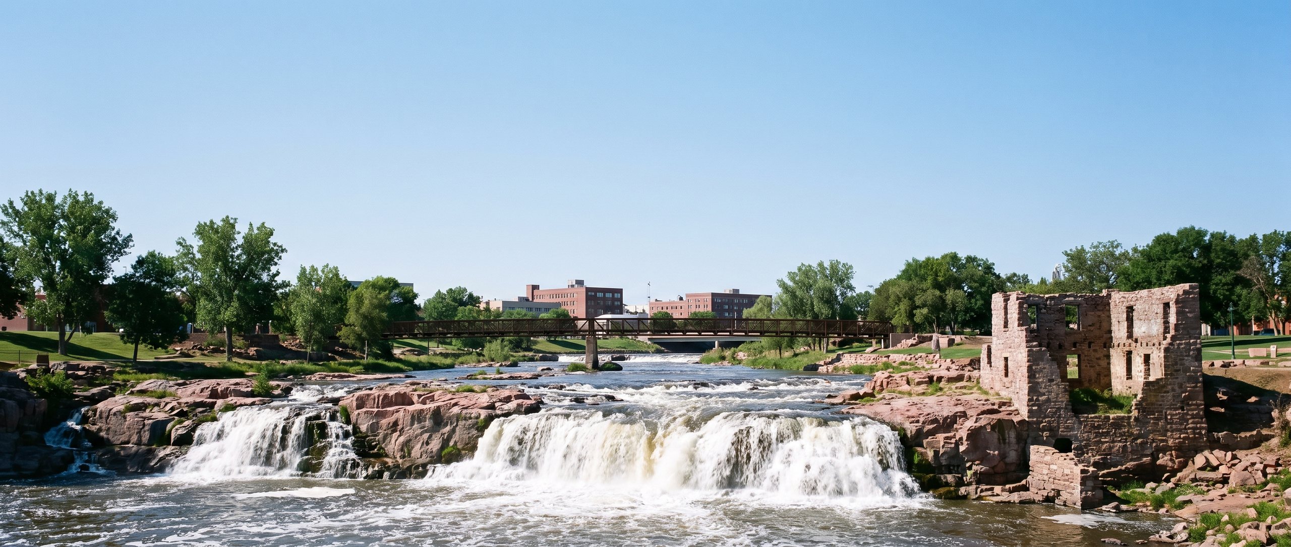 Falls Park in Sioux Falls, South Dakota: the Big Sioux River pouring over pink quartzite ledges, the stone ruins of the old mill on the right, a footbridge and the brick buildings of downtown beyond, under an open sky.